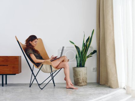 Foto de Mujer sentada en una silla con un ordenador portátil viendo una película y las redes sociales en casa sonrisa, interior moderno y elegante estilo de vida escandinavo, espacio de copia. Foto de alta calidad - Imagen libre de derechos
