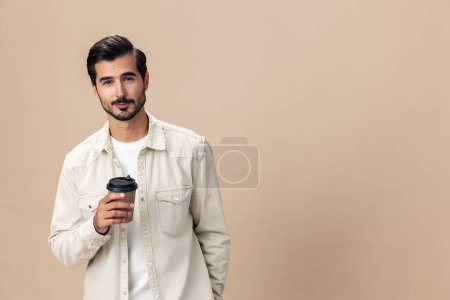 Foto de Retrato de una elegante sonrisa de hombre con una taza de café para ir a burlarse de un fondo beige en una camiseta blanca, estilo de ropa de moda, espacio. Foto de alta calidad - Imagen libre de derechos