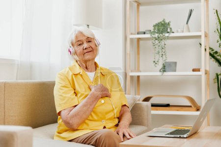 Photo for An elderly woman wearing headphones with a laptop sits on the couch at home and works in a yellow shirt in front of a window. High quality photo - Royalty Free Image