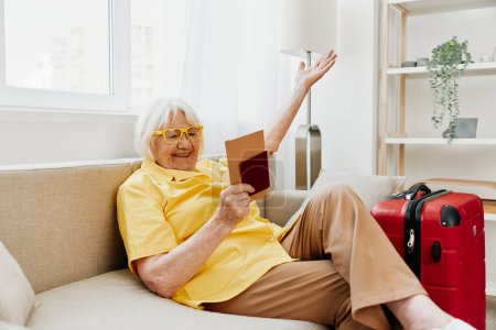 Téléchargez les photos : Heureuse femme âgée avec passeport et billet de voyage emballé une valise rouge, vacances et soins de santé. Une vieille femme souriante s'assoit joyeusement sur le canapé avant le voyage. Photo de haute qualité - en image libre de droit