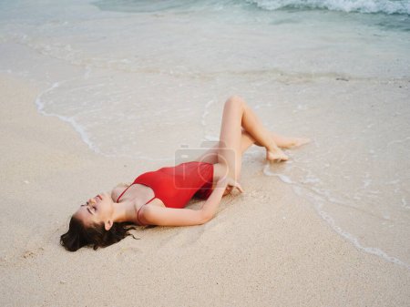 Téléchargez les photos : Une femme en maillot de bain rouge se trouve sur le sable avec un beau corps bronzé du soleil et regarde dans la caméra de l'océan sous les tropiques sur une plage de l'île. Photo de haute qualité - en image libre de droit