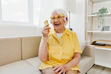 Téléchargez les photos : Heureuse femme âgée regardant dans son appel vidéo téléphonique avec le sourire casque, la technologie pour la communication, intérieur moderne lumineux, mode de vie communication en ligne. Photo de haute qualité - en image libre de droit