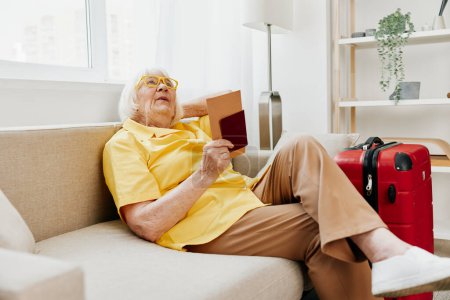 Téléchargez les photos : Heureuse femme âgée avec passeport et billet de voyage emballé une valise rouge, vacances et soins de santé. Une vieille femme souriante s'assoit joyeusement sur le canapé avant le voyage. Photo de haute qualité - en image libre de droit