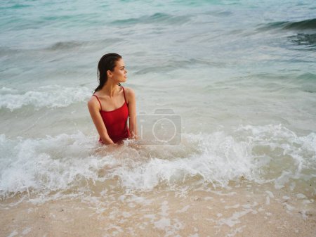 Téléchargez les photos : Femme sexy en maillot de bain rouge avec un corps athlétique se tient dans l'eau dans l'océan et pose en regardant à l'horizon. Photo de haute qualité - en image libre de droit