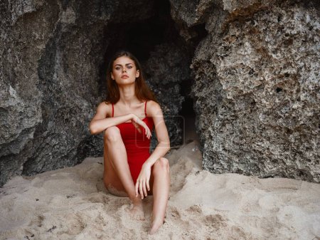 Téléchargez les photos : Femme en maillot de bain rouge assis près des rochers sur le sable avec un beau bronzage du soleil sur la plage, voyage à Bali, des vacances tropicales. Photo de haute qualité - en image libre de droit