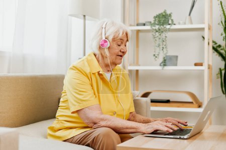 Photo for An elderly woman with headphones and a laptop sits on the couch at home and works. High quality photo - Royalty Free Image