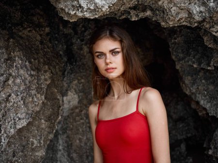 Téléchargez les photos : Portrait d'une femme en maillot de bain rouge contre les rochers au bord de l'océan, peau bronzée du soleil, concept de protection de la peau contre le soleil et santé du corps. Photo de haute qualité - en image libre de droit