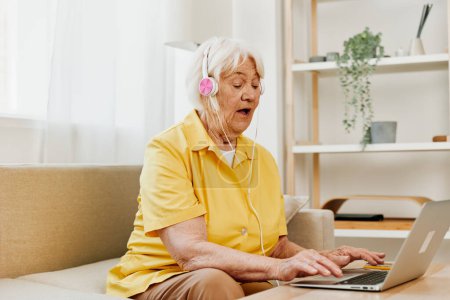 Photo for An elderly woman with headphones and a laptop sits on the couch at home and works. High quality photo - Royalty Free Image