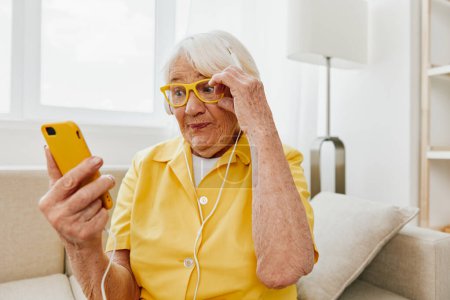 Téléchargez les photos : Heureuse femme âgée regardant un appel vidéo au téléphone et écoutant de la musique sur écouteurs, bouche ouverte et surprise, intérieur moderne lumineux, mode de vie communication en ligne. Photo de haute qualité - en image libre de droit