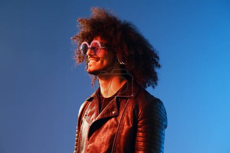 Photo for Portrait of a stylish man with curly hair with glasses and headphones on a blue background multinational, colored light, black leather jacket trend, modern concept. High quality photo - Royalty Free Image
