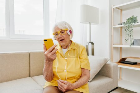 Photo for Happy elderly woman watching video call on phone and listening to music on headphones, surprise and open mouth, bright modern interior, lifestyle online communication. High quality photo - Royalty Free Image