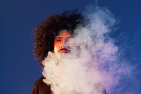 Foto de Retrato de un hombre elegante con el pelo rizado en el humo sobre un fondo azul multinacional, luz de color, tendencia. Foto de alta calidad - Imagen libre de derechos