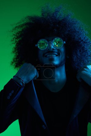 Téléchargez les photos : Portrait de mode d'un homme aux cheveux bouclés sur fond vert avec des lunettes de soleil, multinationale, colorée lumière rose, tendance, concept moderne. Photo de haute qualité - en image libre de droit