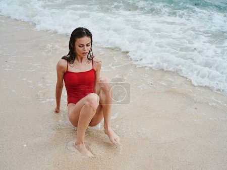 Téléchargez les photos : Femme avec un beau touriste bronzé dans un maillot de bain rouge assis sur le sable sur la plage dans l'océan dans les vagues, Voyage et vacances à la plage. Photo de haute qualité - en image libre de droit