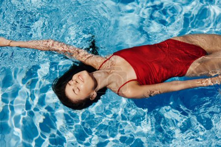 Foto de Mujer joven en la piscina en un traje de baño rojo con una hermosa sonrisa tumbada en el agua y nadando al sol nadando en la piscina, el concepto de relajarse en vacaciones. Foto de alta calidad - Imagen libre de derechos