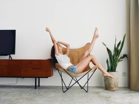 Photo for Woman lying in a leather armchair raised arms and legs cheerful happiness and a smile, relaxing at home stylish modern interior with white walls, copy space. High quality photo - Royalty Free Image