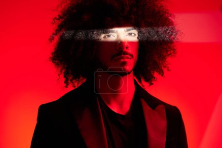 Téléchargez les photos : Portrait de mode d'un homme aux cheveux bouclés sur fond rouge, multinational, lumière colorée, tendance, concept moderne. Photo de haute qualité - en image libre de droit