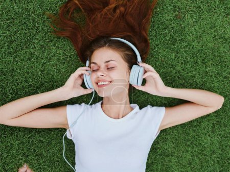Foto de Retrato de mujer tumbada en la hierba verde escuchando música en auriculares con una camiseta blanca, sonrisa con dientes felicidad mirar a la cámara. Foto de alta calidad - Imagen libre de derechos