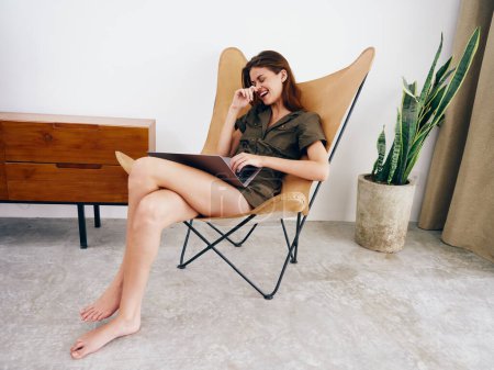 Téléchargez les photos : Femme indépendante assise sur une chaise avec un ordinateur portable à la maison beau sourire et détente, style de vie scandinave intérieur élégant moderne, espace de copie. Photo de haute qualité - en image libre de droit