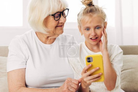 Photo for Two granddaughter smiling photography phone hugging family grandmother selfie indoors people copy call space white child education bonding t-shirt sofa video togetherness - Royalty Free Image