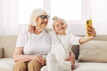 Foto de Selfie blanco niño abrazando abuela dos sonriente teléfono fotografía familia personas sofá llamada unión juntos camiseta nieta espacio vídeo copia educación interior - Imagen libre de derechos