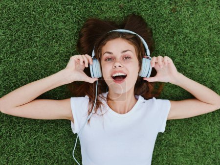 Téléchargez les photos : Une femme heureuse allongée sur le gazon vert dans un T-shirt blanc et écoutant de la musique avec son casque ouvert. Photo de haute qualité - en image libre de droit