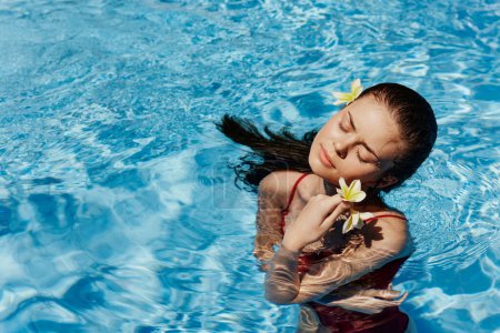 Téléchargez les photos : Jeune femme dans la piscine en maillot de bain rouge avec beau sourire et fleur balinaise au soleil nageant dans la piscine, concept de détente en vacances. Photo de haute qualité - en image libre de droit