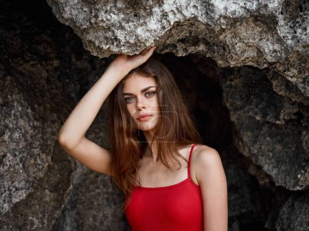 Téléchargez les photos : Portrait d'une femme en maillot de bain rouge contre les rochers au bord de l'océan regardant dans la caméra, peau bronzée du soleil, le concept de protéger la peau du soleil et la santé du corps. Photo de haute qualité - en image libre de droit