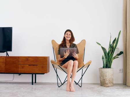 Téléchargez les photos : Étudiante étude s'assoit sur une chaise avec un travail d'ordinateur portable à la maison sourire, style de vie scandinave intérieur élégant moderne, espace de copie. Photo de haute qualité - en image libre de droit