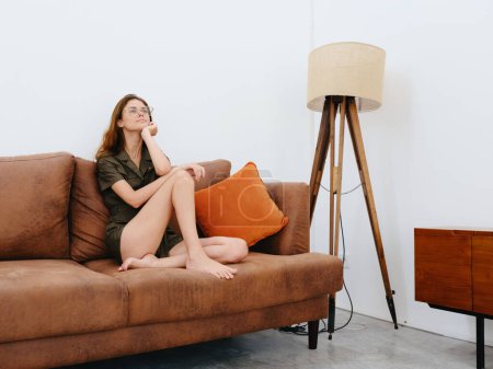 Foto de Modelo de mujer sentada en el sofá en casa sonriente, reflexivo y relajado, estilo de vida interior moderno, espacio para copiar, paleta de colores de otoño. Foto de alta calidad - Imagen libre de derechos