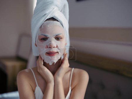 Téléchargez les photos : Jeune femme couchée sur le lit avec masque hydratant beauté et serviette blanche sur sa tête après la douche, mode de vie soins de la peau à la maison, sourire. Photo de haute qualité - en image libre de droit