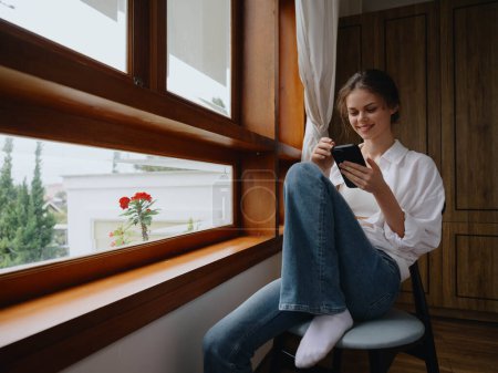 Foto de Hermosa mujer con teléfono en la mano sentada en la ventana con marco de madera de la casa, hogar cómodo estilo de vida con trabajo en línea, ambiente acogedor y estética, la primavera. Foto de alta calidad - Imagen libre de derechos