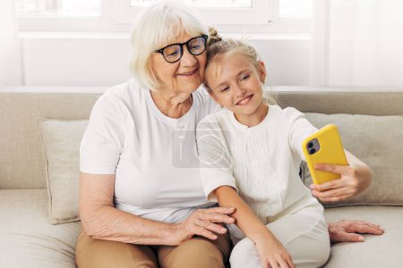 Photo for Space two sofa granddaughter hugging phone t-shirt photography white grandmother people selfie child bonding togetherness family video education call copy smiling indoors - Royalty Free Image