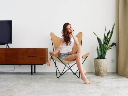 Photo for Woman sitting in leather armchair smile with teeth Lifestyle in white T-shirt and blue denim shorts, resting at home stylish modern interior with white walls, copy space. High quality photo - Royalty Free Image