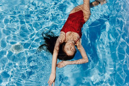Foto de Mujer joven en la piscina en traje de baño rojo con una hermosa sonrisa acostada en el agua y nadando en la piscina azul en el sol, el concepto de relajarse en vacaciones. Foto de alta calidad - Imagen libre de derechos