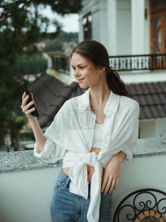 Téléchargez les photos : Femme sur le balcon de la maison avec téléphone beau sourire un jour de congé, belle vue sur la nature dans les montagnes, vacances style de vie dans l'hôtel. Photo de haute qualité - en image libre de droit