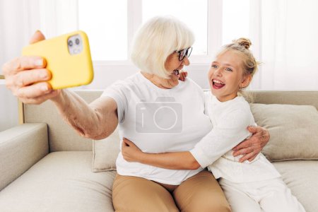 Téléchargez les photos : Grand-mère enfant selfie canapé collage t-shirt copie bonheur ensemble vidéo téléphone éducation photographie étreinte famille gens deux espace intérieur petite-fille appeler blanc sourire - en image libre de droit