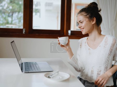 Téléchargez les photos : Jeune femme créative travaillant à un bureau à la maison dans un intérieur confortable avec une tasse de boisson, bureau à domicile pigiste avec un sourire, la communication et l'éducation en ligne. Photo de haute qualité - en image libre de droit