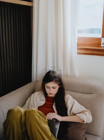 Téléchargez les photos : Une femme s'allonge sur un canapé avec un ordinateur portable et regarde pensivement à l'écran former un pigiste au travail, un vrai mode de vie sans retoucher. Photo de haute qualité - en image libre de droit