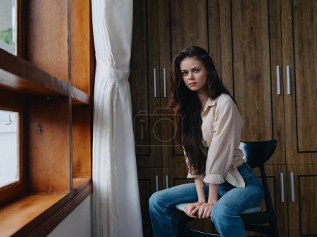Foto de Mujer sentada en una silla en el interior del hogar cerca de la ventana, comodidad y comodidad, relajación estilo de vida en el fin de semana, aspecto pensativo de una hermosa chica. Foto de alta calidad - Imagen libre de derechos