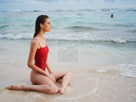 Téléchargez les photos : Une jeune femme en maillot de bain rouge est assise sur le sable avec un beau bronzage et regarde l'océan sous les tropiques sur une plage insulaire. Photo de haute qualité - en image libre de droit