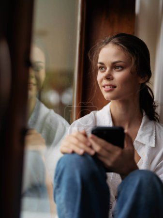 Téléchargez les photos : Jeune femme assise sur un rebord de fenêtre en bois près de la fenêtre avec un téléphone à la main souriant et regardant par la fenêtre, humeur d'automne, atmosphère confortable à la maison, style de vie esthétique. Photo de haute qualité - en image libre de droit