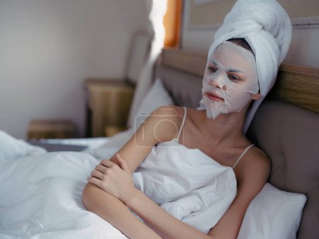 Foto de Mujer joven acostada en la cama con mascarilla hidratante belleza y toalla blanca en la cabeza después de la ducha, estilo de vida cuidado de la piel en casa, sonrisa. Foto de alta calidad - Imagen libre de derechos