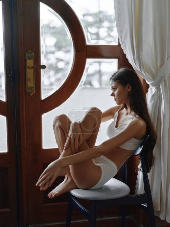 Photo for Young slim brunette woman sitting on a chair by the window of the house in comfortable home linen, summer atmosphere, lifestyle loneliness. High quality photo - Royalty Free Image