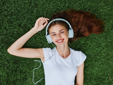 Foto de Una hermosa mujer con auriculares escucha música tumbada en el césped del parque en primavera y sonríe. Foto de alta calidad - Imagen libre de derechos