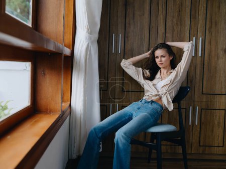 Téléchargez les photos : Femme assise sur une chaise à l'intérieur de la maison près de la fenêtre, confort et confort, détente de style de vie le week-end, regard cher d'une belle fille. Photo de haute qualité - en image libre de droit