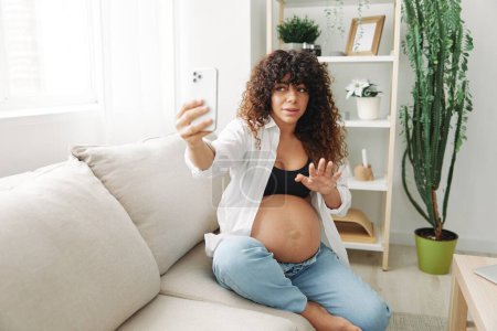 Foto de La bloguera embarazada se sienta en el sofá de su casa y se toma fotos por teléfono, selfies y videollamadas, consulta con el médico en línea, manejo del embarazo. Foto de alta calidad - Imagen libre de derechos