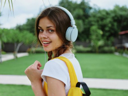 Téléchargez les photos : Heureuse adolescente écoutant de la musique avec un casque avec un sac à dos dans un T-shirt blanc et marchant jusqu'à l'université pour étudier dans la rue en souriant avec des dents, mode de vie étudiant. Photo de haute qualité - en image libre de droit