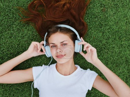 Téléchargez les photos : Une femme allongée sur la pelouse verte écoutant de la musique dans un casque en T-shirt blanc, un sourire avec des dents de bonheur. Photo de haute qualité - en image libre de droit