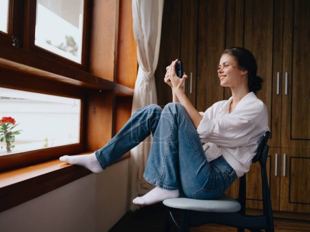 Foto de Mujer joven sentada junto a la ventana con la sonrisa del teléfono en la mano buscando en línea, el estado de ánimo de primavera, ropa sencilla en casa, ambiente acogedor en casa, estilo de vida estético. Foto de alta calidad - Imagen libre de derechos